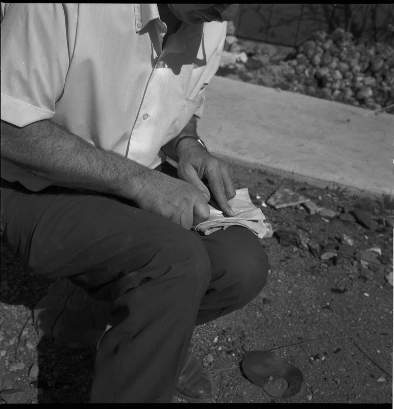 An image of Donald E. Crabtree flintknapping on a log stump. Crabtree is using a possibly bone tool to pressure flake the edge he just prepared by grounding it with the hammerstone.