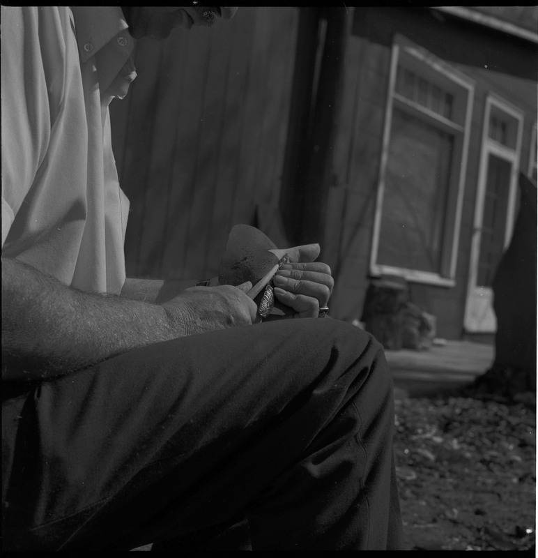 An image of Donald E. Crabtree flintknapping on a log stump. Crabtree is holding the stone object he is working with. He is holding it flat on the palm of his left hand, covered with a piece of leather, while he uses a bone tool to pressure flake the object.