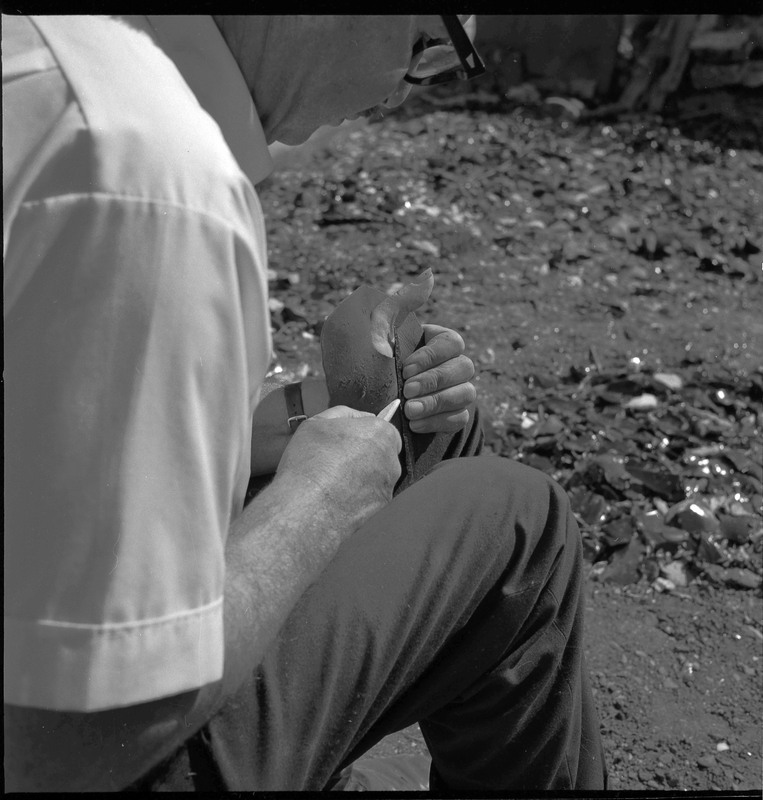 An image of Donald E. Crabtree flintknapping on a log stump. Crabtree is holding the stone object he is working with. He is holding it flat on the palm of his left hand, covered with a piece of leather, while he uses a bone tool to pressure flake the object.