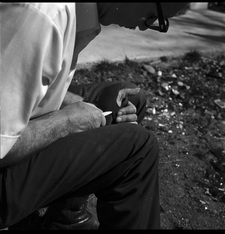 An image of Donald E. Crabtree flintknapping on a log stump. Crabtree is holding the stone object he is working with. Working on the object, he is holding it flat on the palm of his left hand, covered with a piece of leather, while he uses a bone tool to pressure flake the object.
