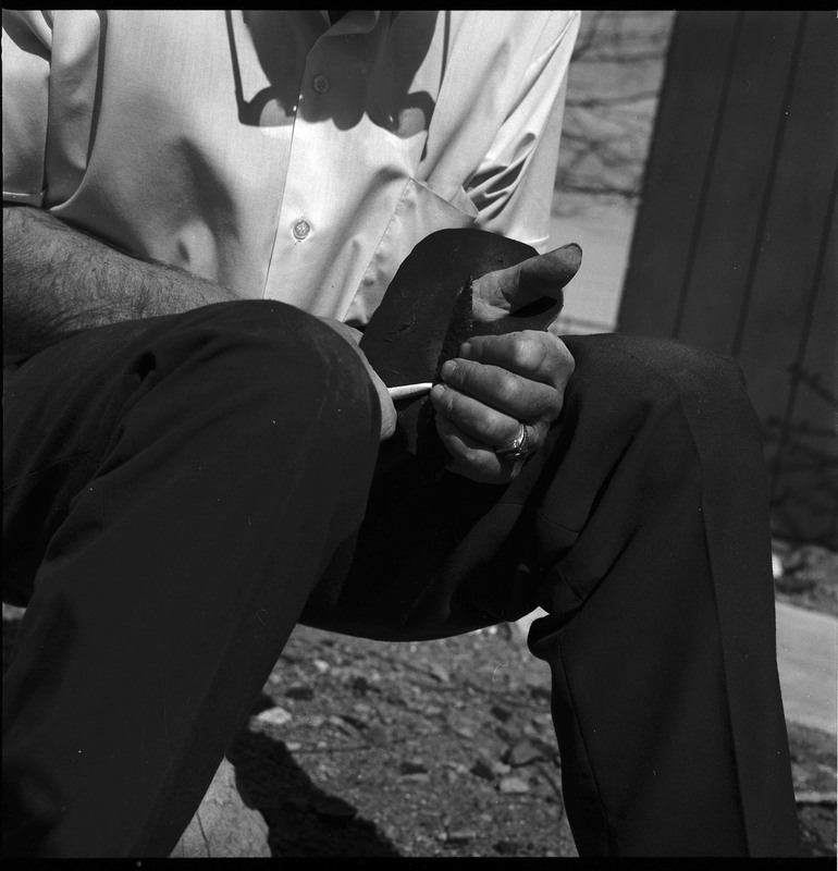 An image of Donald E. Crabtree flintknapping on a log stump. Crabtree is holding the stone object he is working with. Working on the object, he is holding it flat on the palm of his left hand, covered with a piece of leather, while he uses a bone tool to pressure flake the object.