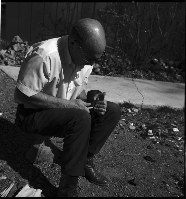 An image of Donald E. Crabtree flintknapping on a log stump. Crabtree is holding the stone object he is working with. Working on the object, he is holding it flat on the palm of his left hand, covered with a piece of leather, while he uses a bone tool to pressure flake the object.