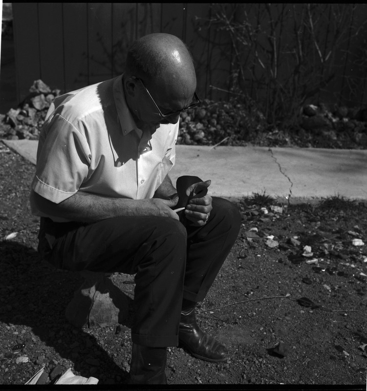 An image of Donald E. Crabtree flintknapping on a log stump. Crabtree is holding the stone object he is working with. Working on the object, he is holding it flat on the palm of his left hand, covered with a piece of leather, while he uses a bone tool to pressure flake the object.