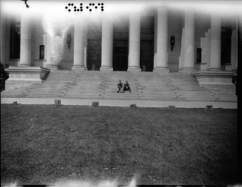 A black and white negative of two people sitting side by side on the stairs of a large building. The building has columns and is detailed. The photo is taken from far away with grass in the frame.