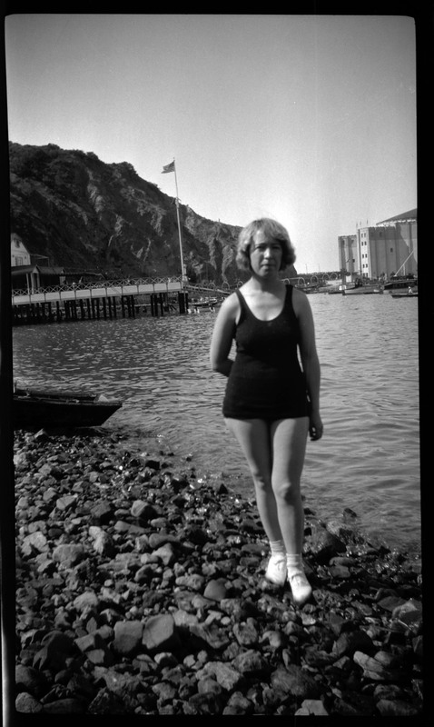 A black and white negative of a woman in a bathing suit standing on a rocky shoreline of a body of water. There is a mountain, dock, large building, and boat in the background. There is an American flag in the photo. Likely on Catalina island.
