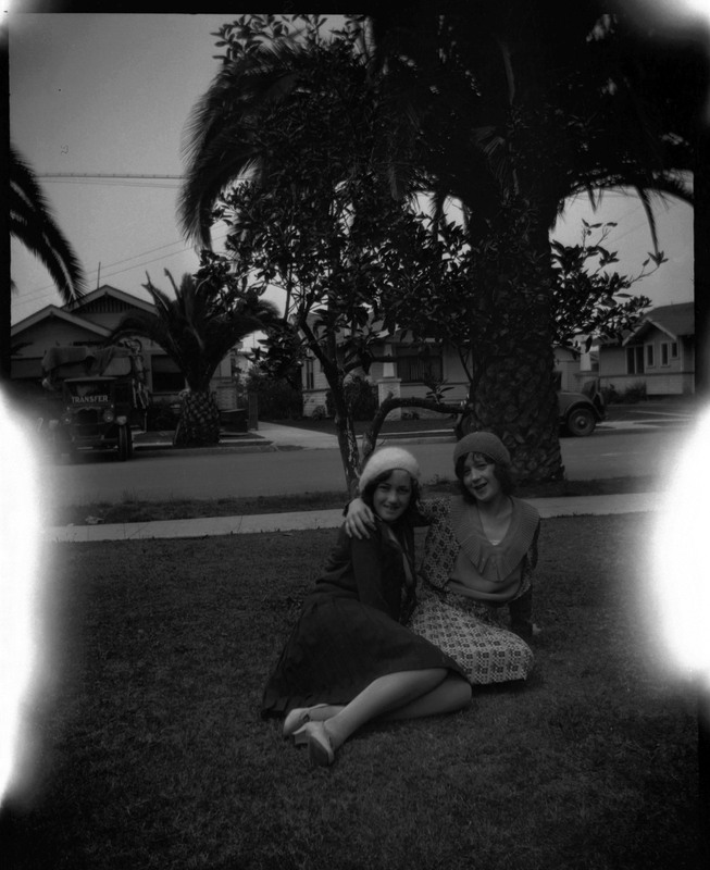 A black and white negative of young Evelyn Crabtree and another woman arm in arm sitting on a lawn. They are sitting in a house's front yard, there are large palm trees in the background, and a moving truck across the street.