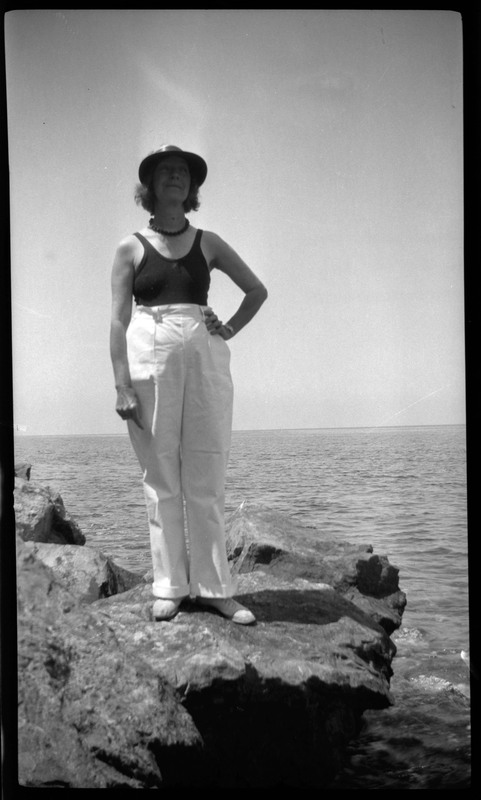 A black and white negative of a woman standing on a rock by the ocean. There is vast ocean in the background. The woman is wearing a bathing suit, a hat, and pants.
