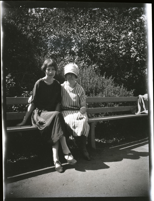 A black and white negative of two women sitting on a bench in an outdoor park area. the women are wearing dresses and one is wearing a hat.