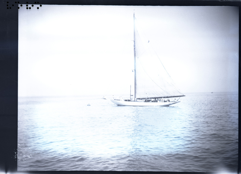 A black and white negative of a sailboat on the ocean. The boat is long and there seems to be two shapes in the water. The boat has people on it.