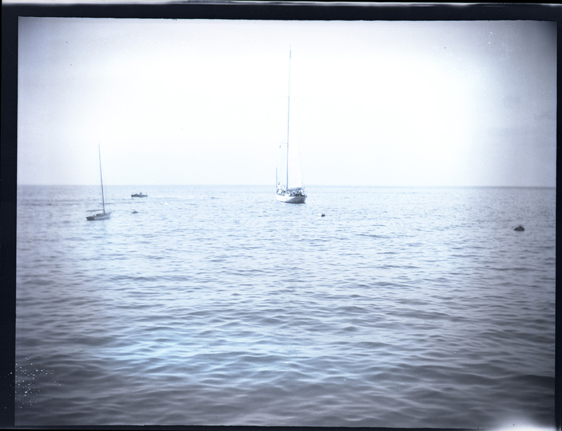 A black and white negative of three sailboats on the ocean. There are three buoys in the water. One boat is far larger than the others.