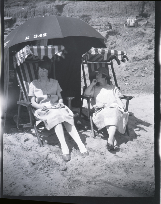 A black and white negative of two women sitting under striped umbrellas on the beach. There are bikes in the background on a rock formation.