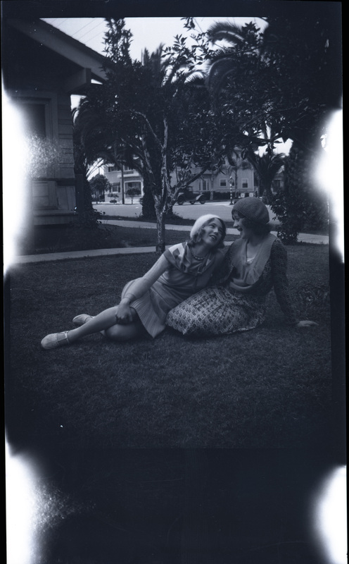 A black and white negative of two women sitting together laughing on a lawn out front of a house. The girls are in dresses and there are palm trees and moving car in the background.