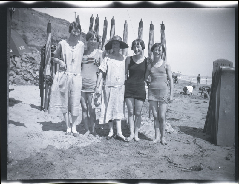 A black and white negative of five women in bathing suits standing together on the beach. There are folded umbrellas behind them and people on the coastline.