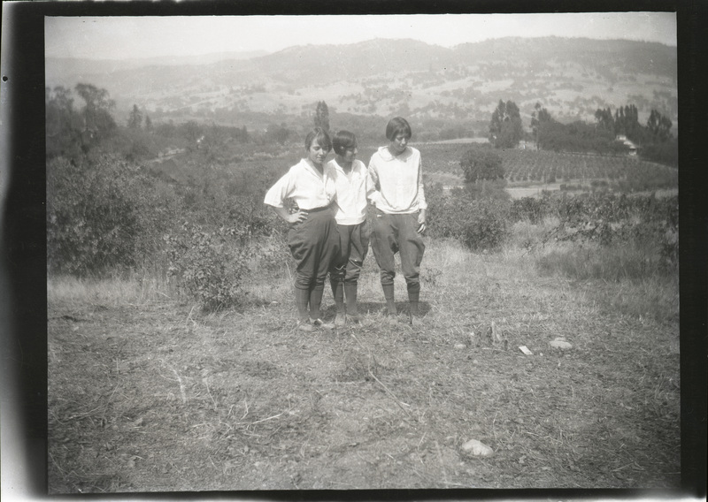 A black and white negative of three women standing together on a hilltop. There seems to be a vineyard or orchard behind them. There is a large hilly background.