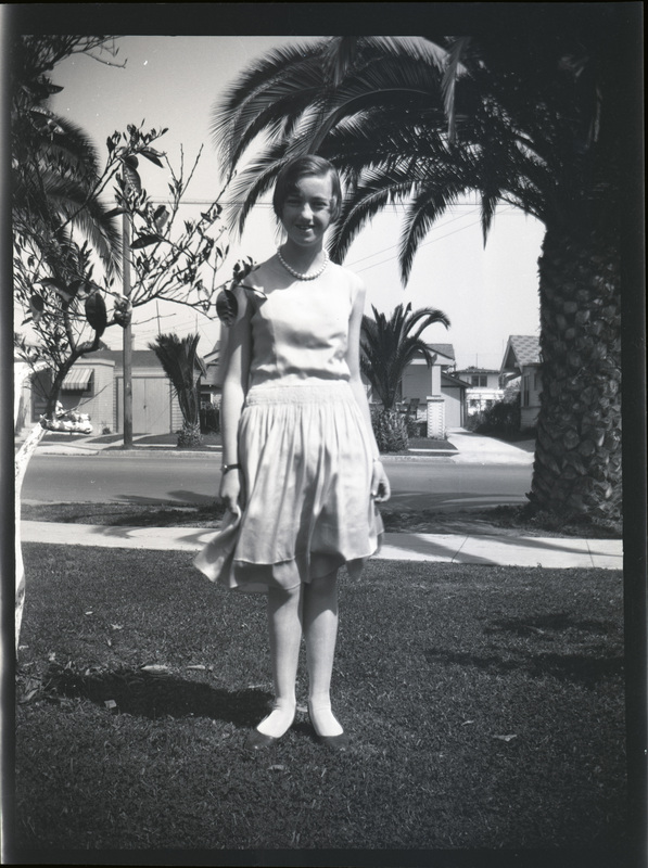 A black and white negative of Evelyn Crabtree as young girl. She is standing on her front lawn in a nice dress and pearls. There are palm trees and a neighborhood in the background.
