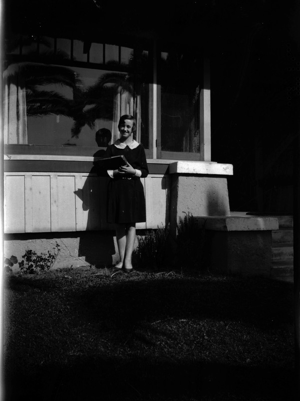 A black and white negative of Evelyn Crabtree as a young schoolgirl with books. She is standing in front of a house.