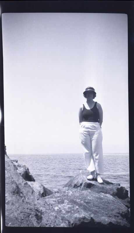 A black and white negative of a woman standing in front of the ocean on a large rock. She is wearing a swimsuit with a sunhat.