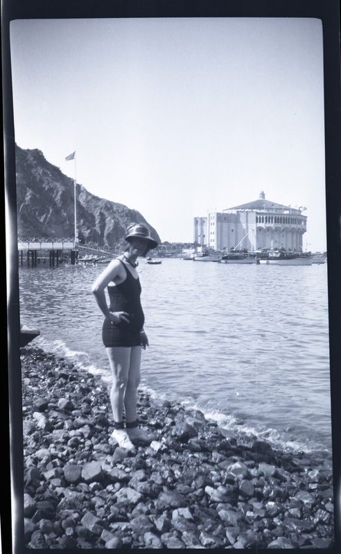 A black and white negative of a woman in a bathing suit standing on a rocky shoreline of the ocean. There is a large hotel looking building in the background, boats, a dock, and an American flag. Likely on Catalina island.