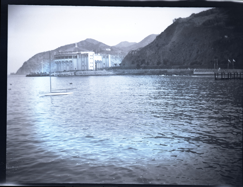 A black and white negative of the ocean and shoreline of Catalina Casino on Catalina Island of California. There are boats in the photo.