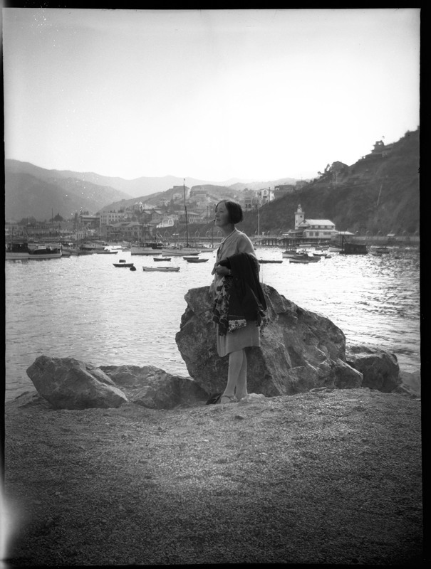 A black and white negative of a woman standing on a rock by the ocean in front of sailboats and buildings. Likely on Catalina Island.