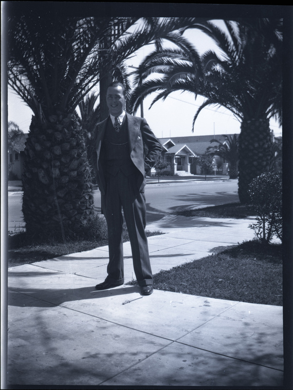 A black and white negative of a young man in a nice suit standing in a driveway. There are palm trees in the background and other houses.