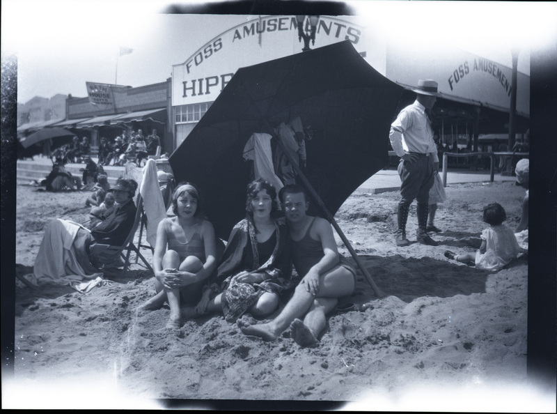 A black and white negative of three young people sitting under an umbrella on the beach. There are other people in the background and beach toursit buildings behind them.