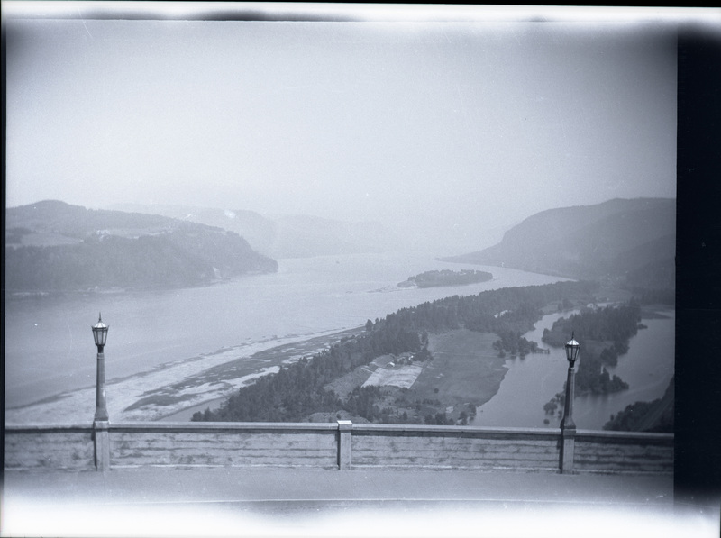 A black and white negative of a river canyon from a bridge or road. There are hills and trees along the coastline.