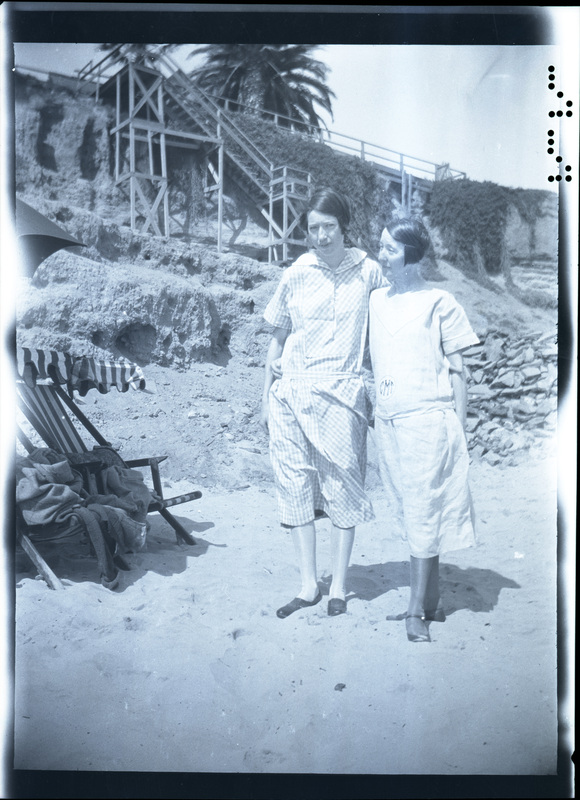 A black and white negative of two women standing together on the beach. There is a large staircase and beach chairs in the background. The women are wearing frocks.