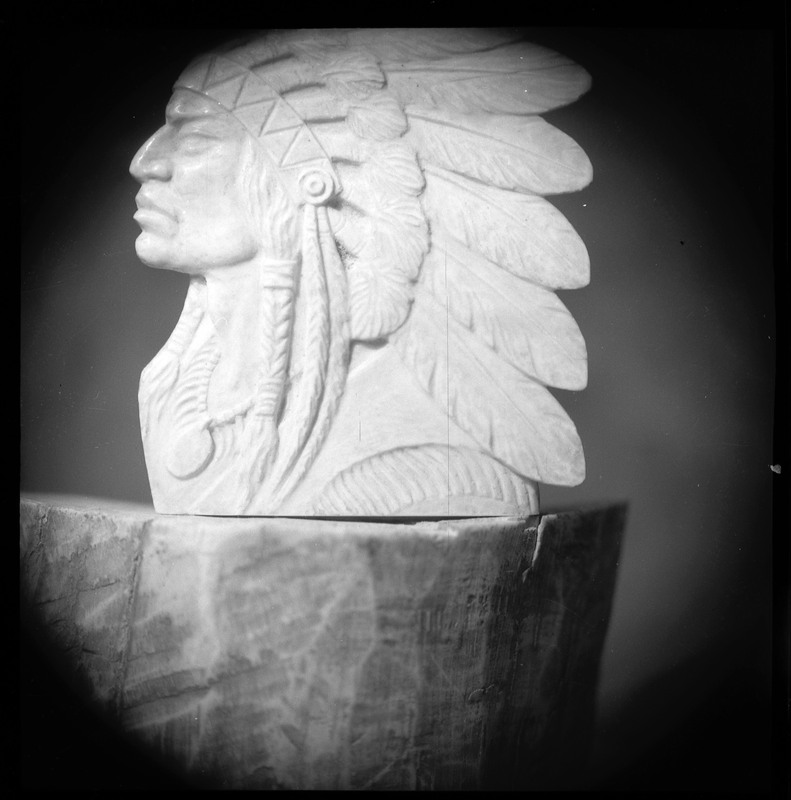 A black and white negative of a carving into stone of an Indigenous man wearing a feathered headdress. The carving is sitting on a log.
