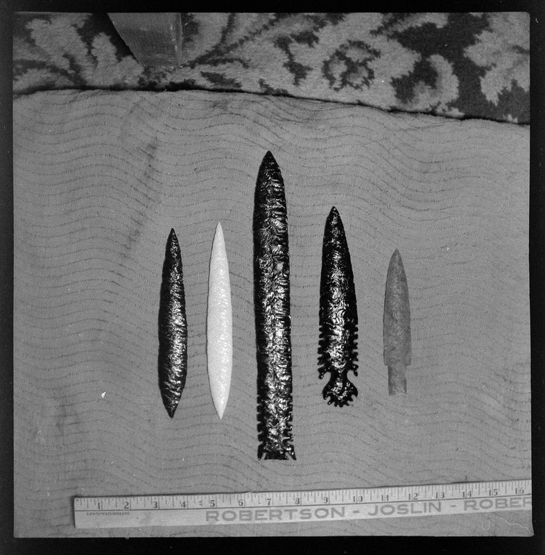 A black and white negative of five bifaces with a yardstick in the photo for reference. There are three black bifaces, one white, and one grey looking one, dependent on the coloring of the photo.