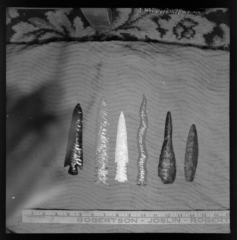 A black and white negative of six bifaces with a yardstick in the photo for reference. This image is obstructed by something on the left side. The bifaces are all different colors and appear to vary in material type. It is difficult to distingush this with the coloring.