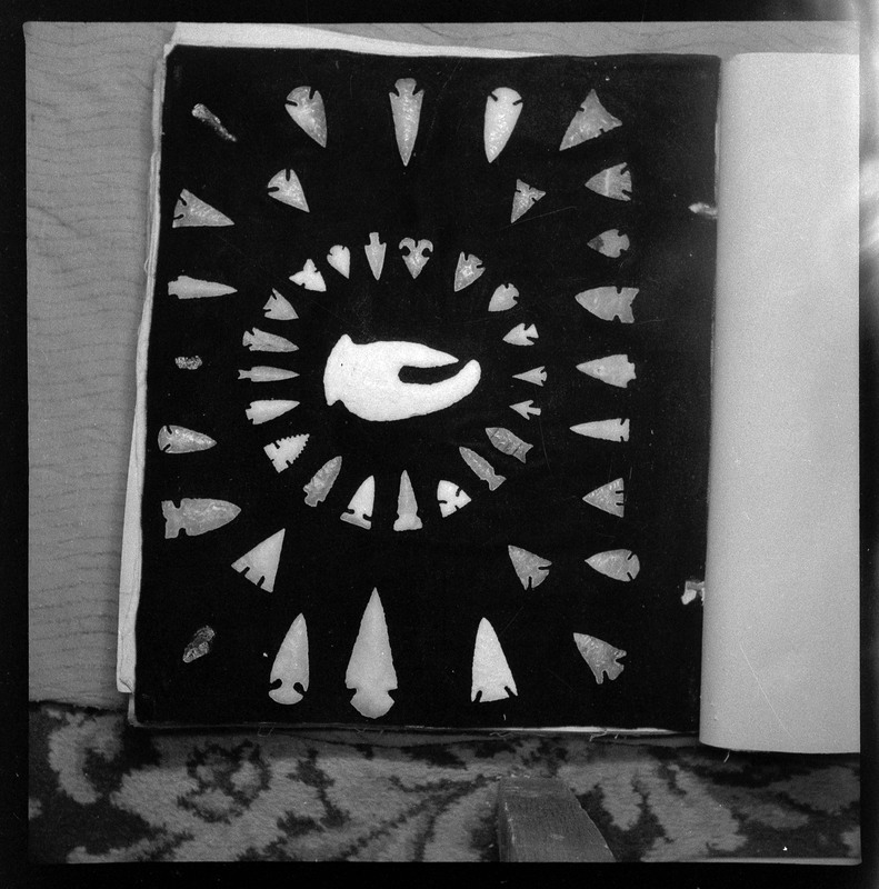 A black and white negative with many bifaces, varying in size, color and style organized in a circle around a large central biface that looks similar to a crab claw.