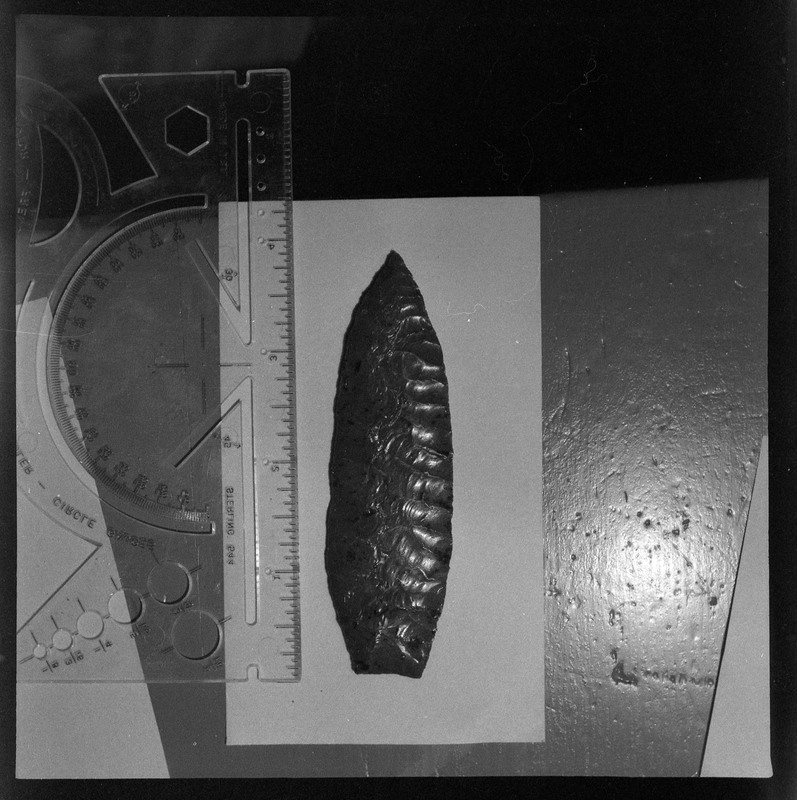 A black and white negative of a close-up view of a biface. The object looks to be obsidian and has a small ruler placed next to it to indiciate size.