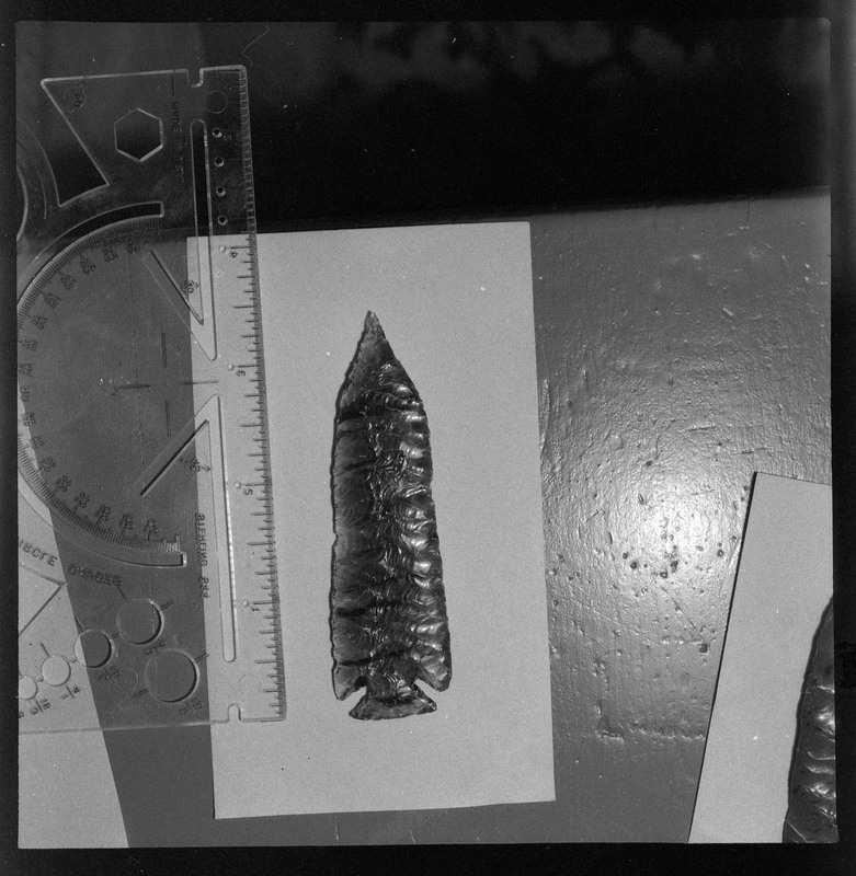 A black and white negative of a close-up view of a biface. The object looks to be obsidian and has a small ruler placed next to it to indiciate size.