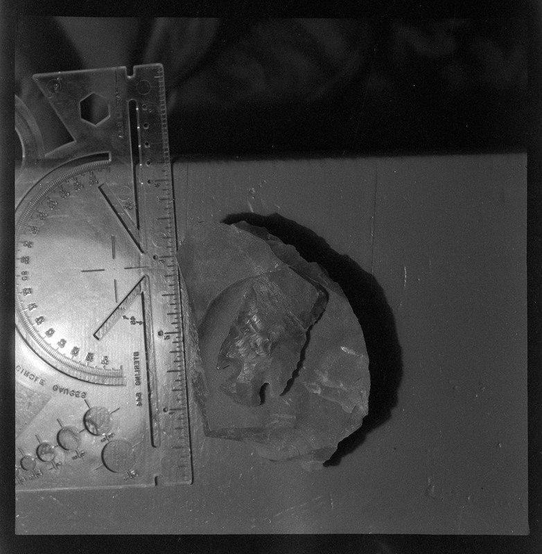 A black and white negative of a close-up view of a broken biface on top of a rock. The material is hard to determine based on photo coloring and has a small ruler placed next to it to indiciate size.