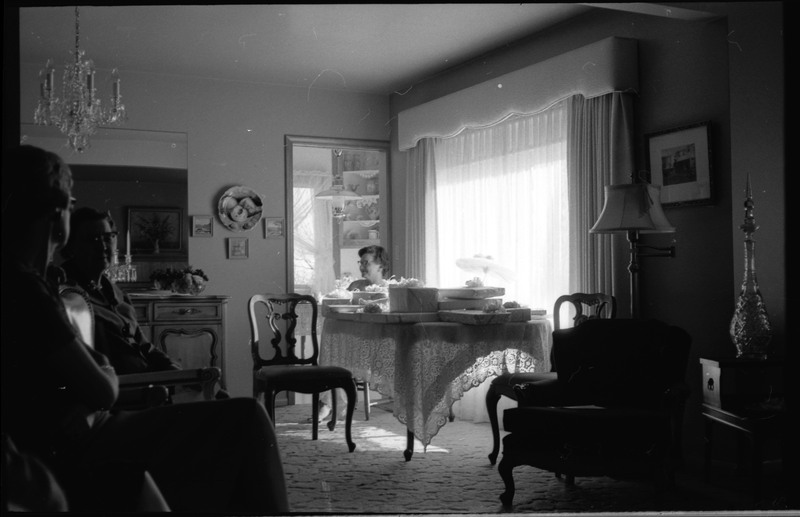 A photo of three people gathered in a sitting room, with two people in the foreground and one woman in the background. The woman in the background is sitting at a table with gifts. The event appears to be a bridal shower. Associated with item numbers: ce_b83_f1-item1-001 through ce_b83_f1-item1-016.