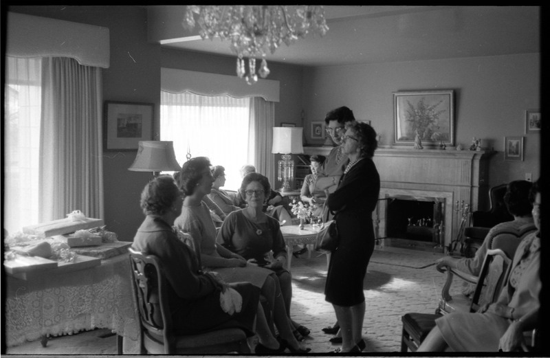 A photo of a group of women gathered in a sitting room. A table of gifts is visible on the left side of the image. The event appears to be a bridal shower. Associated with item numbers: ce_b83_f1-item1-001 through ce_b83_f1-item1-016.