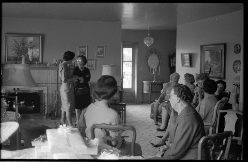 A photo of a group of women gathered in a sitting room. A table of gifts is visible in the foreground of the image. The event appears to be a bridal shower. Associated with item numbers: ce_b83_f1-item1-001 through ce_b83_f1-item1-016.