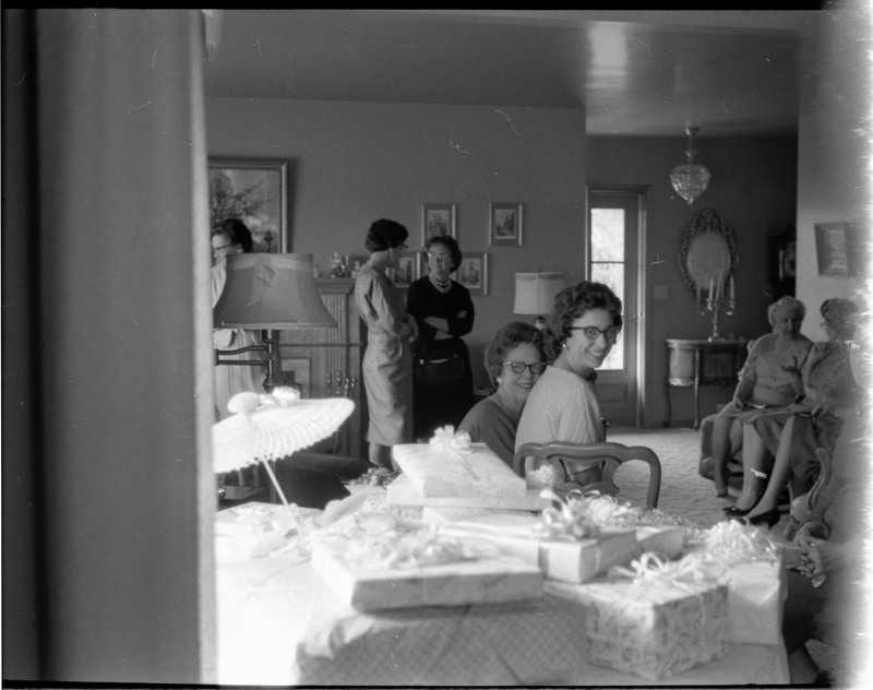 A photo of a group of women gathered in a sitting room. A table of gifts is visible in the foreground of the image and two women are smiling, looking directly at the camera. The event appears to be a bridal shower. Associated with item numbers: ce_b83_f1-item1-001 through ce_b83_f1-item1-016.