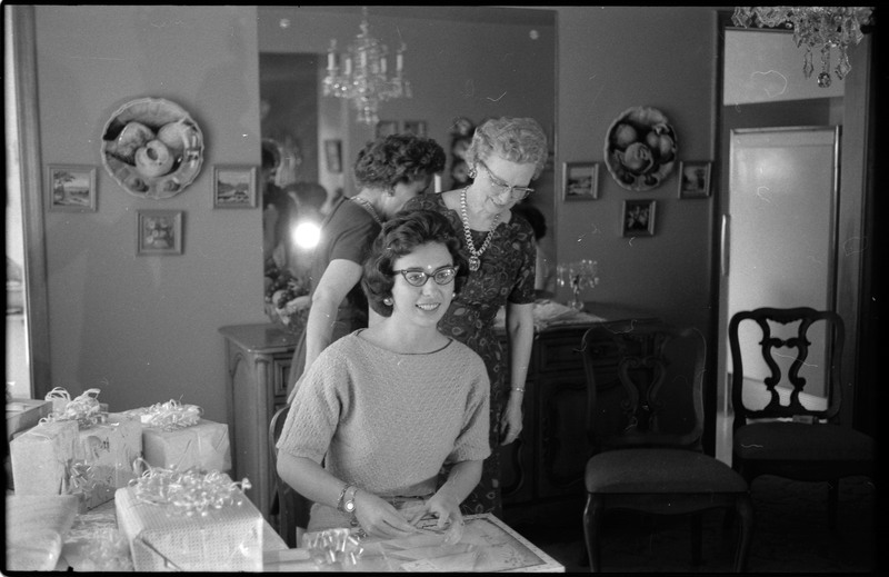 A photo of a woman opening gifts in a sitting room. The event appears to be a bridal shower. Associated with item numbers: ce_b83_f1-item1-001 through ce_b83_f1-item1-016.