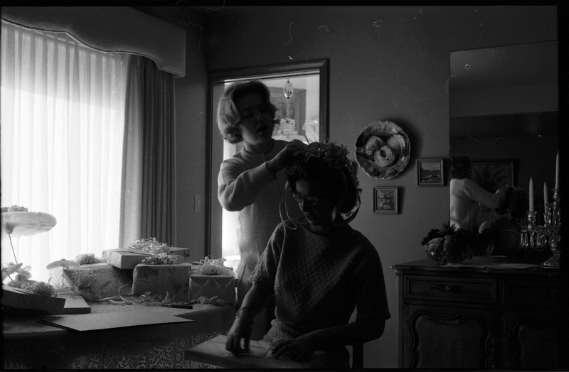 A photo of a woman opening gifts in a sitting room. Another woman stands behing her and appears to be placing bows and ribbons in her hair. The event appears to be a bridal shower. Associated with item numbers: ce_b83_f1-item1-001 through ce_b83_f1-item1-016.