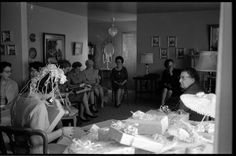 A photo of a woman opening gifts in a sitting room. She is surrounded by a group of other women. The event appears to be a bridal shower. Associated with item numbers: ce_b83_f1-item1-001 through ce_b83_f1-item1-016.