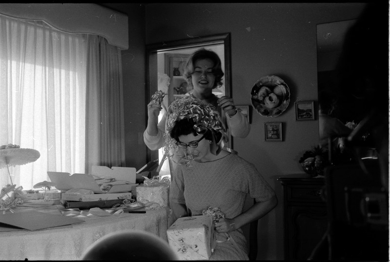 A photo of a woman opening gifts in a sitting room. Another woman stands behing her and appears to be placing bows and ribbons in her hair. The event appears to be a bridal shower. Associated with item numbers: ce_b83_f1-item1-001 through ce_b83_f1-item1-016.