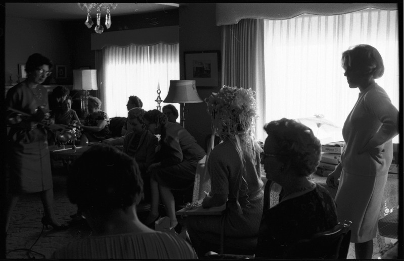 A photo of a group of women gathered in a sitting room. The woman sitting in the center foreground is holding a gift and has a hat of ribbons and bows in her hair. The event appears to be a bridal shower. Associated with item numbers: ce_b83_f1-item1-001 through ce_b83_f1-item1-016.