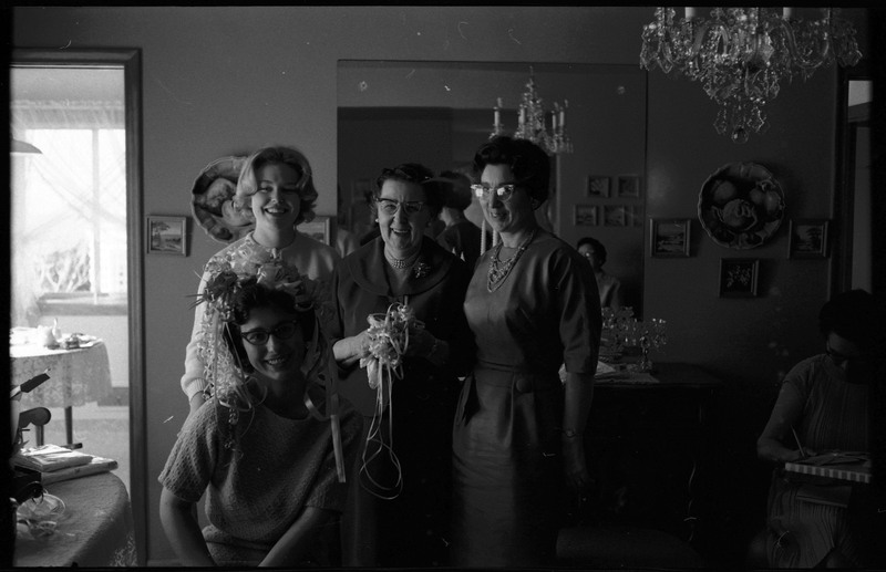 A photo of four women posing for the camera in a sitting room. The seated woman has a hat of of ribbons and bows in her hair. The event appears to be a bridal shower. Associated with item numbers: ce_b83_f1-item1-001 through ce_b83_f1-item1-016.