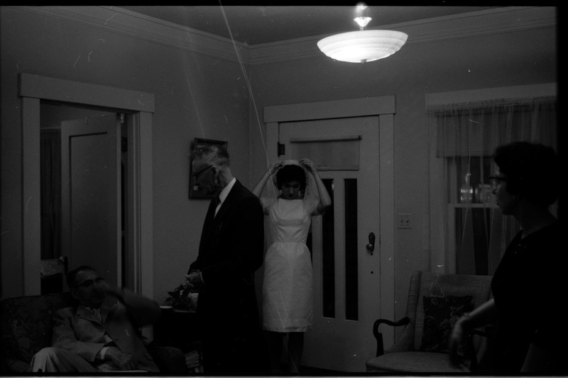 A photo of a woman wearing a wedding dress and adjusting her veil in a room with others. Associated with item numbers: ce_b83_f1-item1-017 through ce_b83_f1-item1-036.