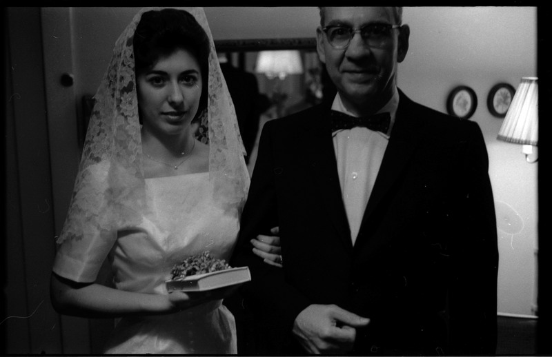 A photo of a woman wearing a wedding dress and a man wearing a suit posing for the camera. The woman is carrying a book. Associated with item numbers: ce_b83_f1-item1-017 through ce_b83_f1-item1-036.