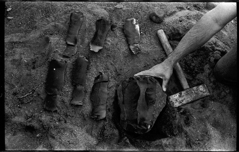 A photo of six flintknapped pieces and a large piece of stone in dirt. A tool, which appears to be an axe, is also visible.