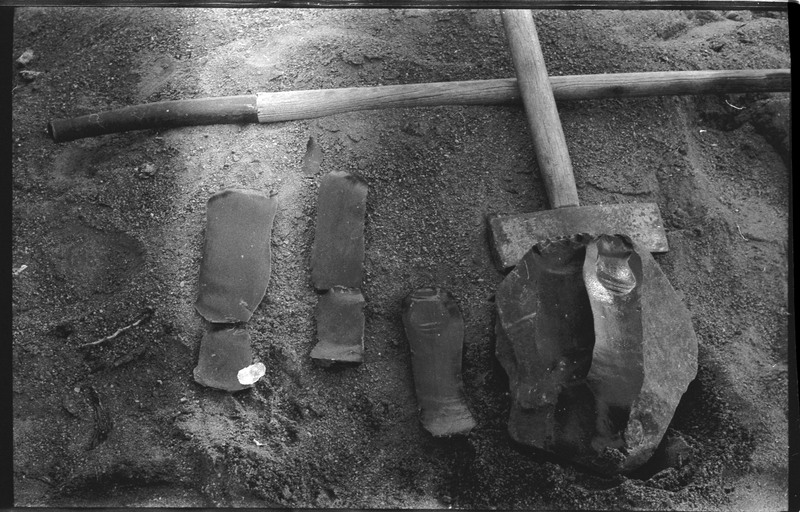 A photo of four flintknapped pieces and a large piece of stone in dirt. Two tools, one of which appears to be an axe, are also visible.