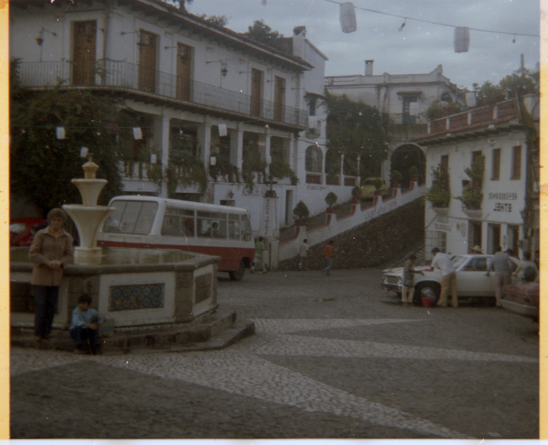 A photograph of Evelyn Crabtree posing next to a fountain in a city square. A young, unnamed child, is sitting in front of the fountain.